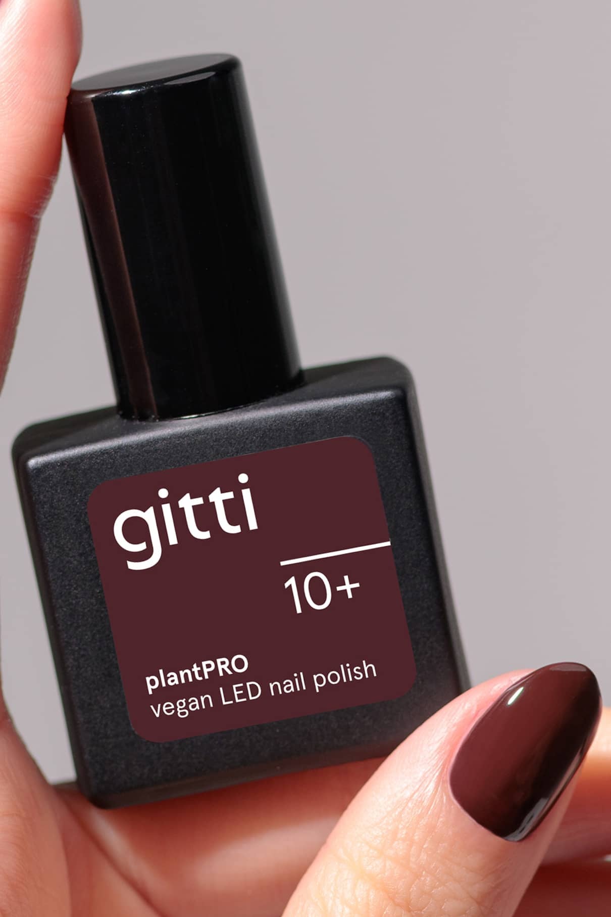 Eine Hand hält eine quadratische Flasche gitti no. 10+ veganer LED-Nagellack in einem dunklen Beerenton, mit passend lackierten Nägeln.