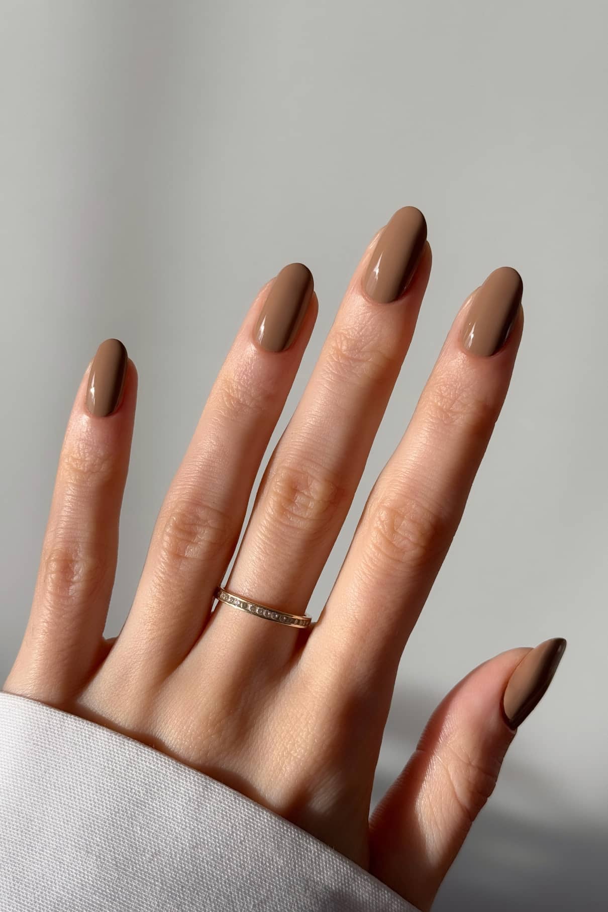 Eine Hand mit mandelförmigen Nägeln, die in gitti no. 09+, einem glänzenden hellen Braunton mit warmen Untertönen, trägt einen schlichten Goldring am Ringfinger vor einem neutralen Hintergrund.