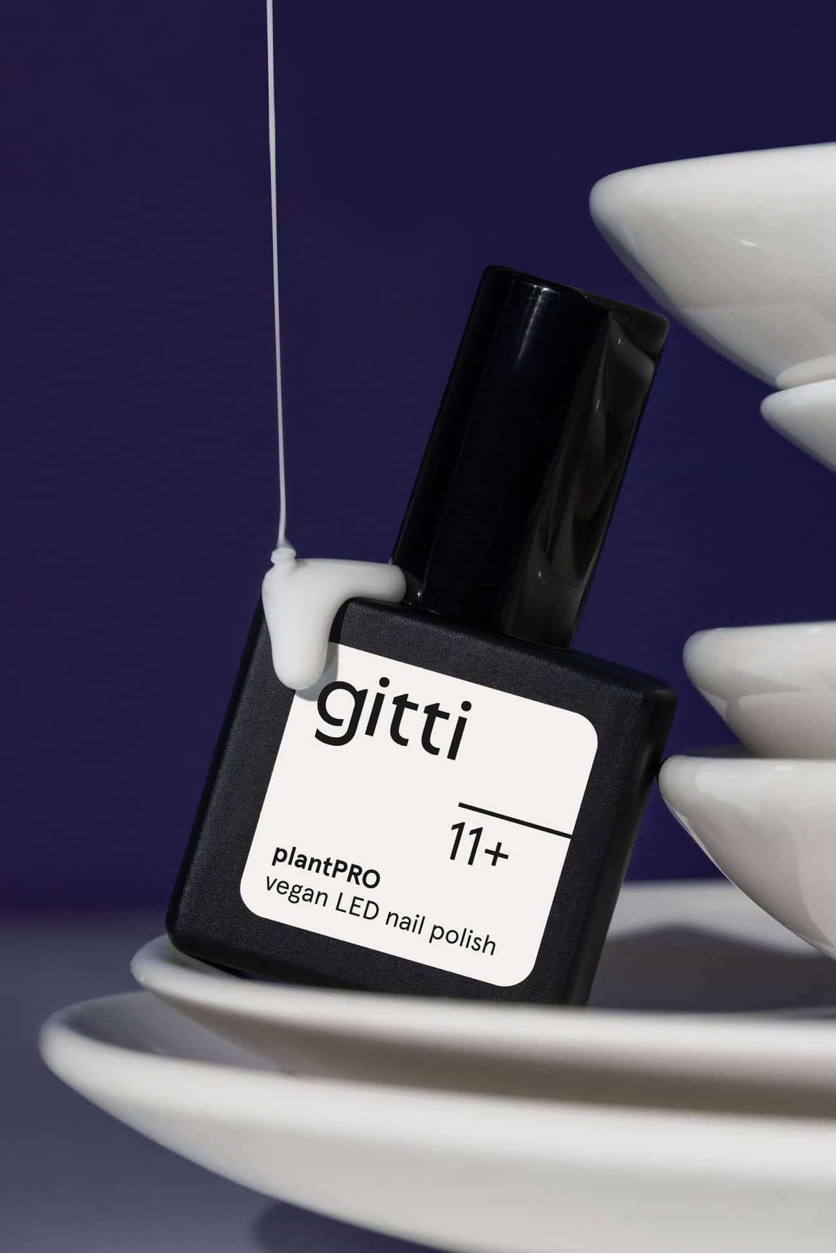 Eine schwarze Flasche mit gitti no. 11+ plantPRO veganer LED-Nagellack in zartes Weiß ruht auf gestapelten weißen Tellern, über die sich eine weiße Flüssigkeit ergießt - ein Look, der zu halbtransparentem Nageldesign inspiriert.
