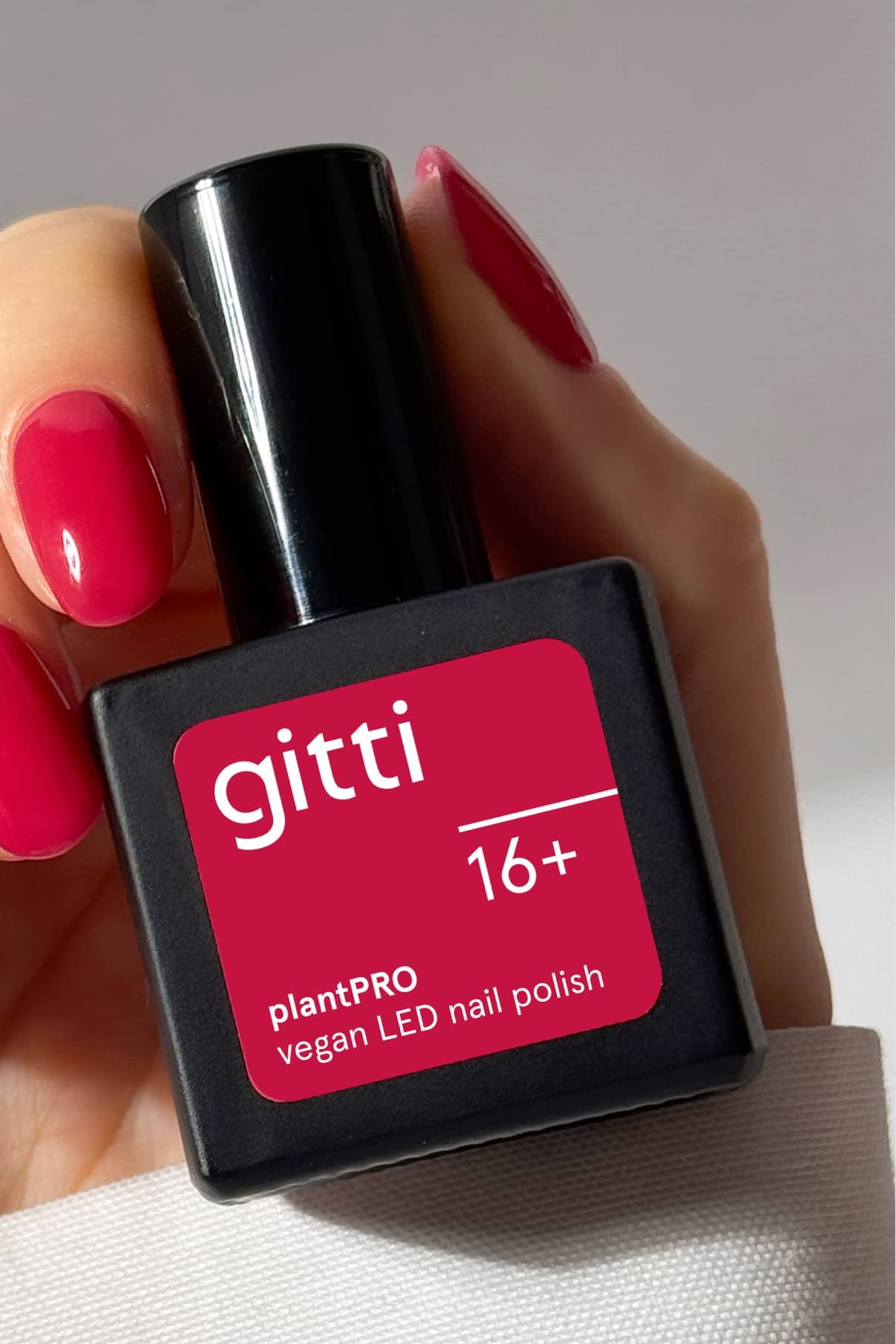 Eine Hand mit intensiv rosa lackierten Nägeln hält ein quadratisches, schwarzes Fläschchen mit der Aufschrift "gitti no. 16+".