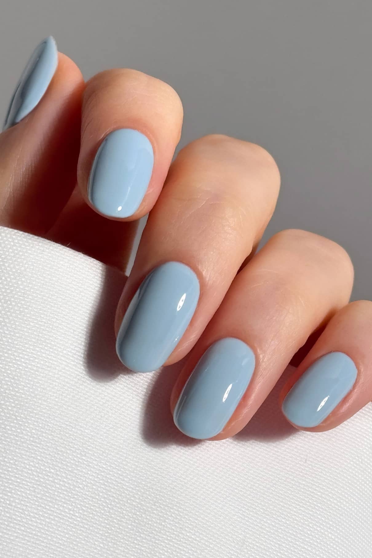 Eine Hand mit sauber manikürten Nägeln, die in einem glänzenden hellblauen PRO Colour Duo lackiert und mit einem glatten Decklack versehen sind, ruht auf einer weißen, strukturierten Oberfläche.