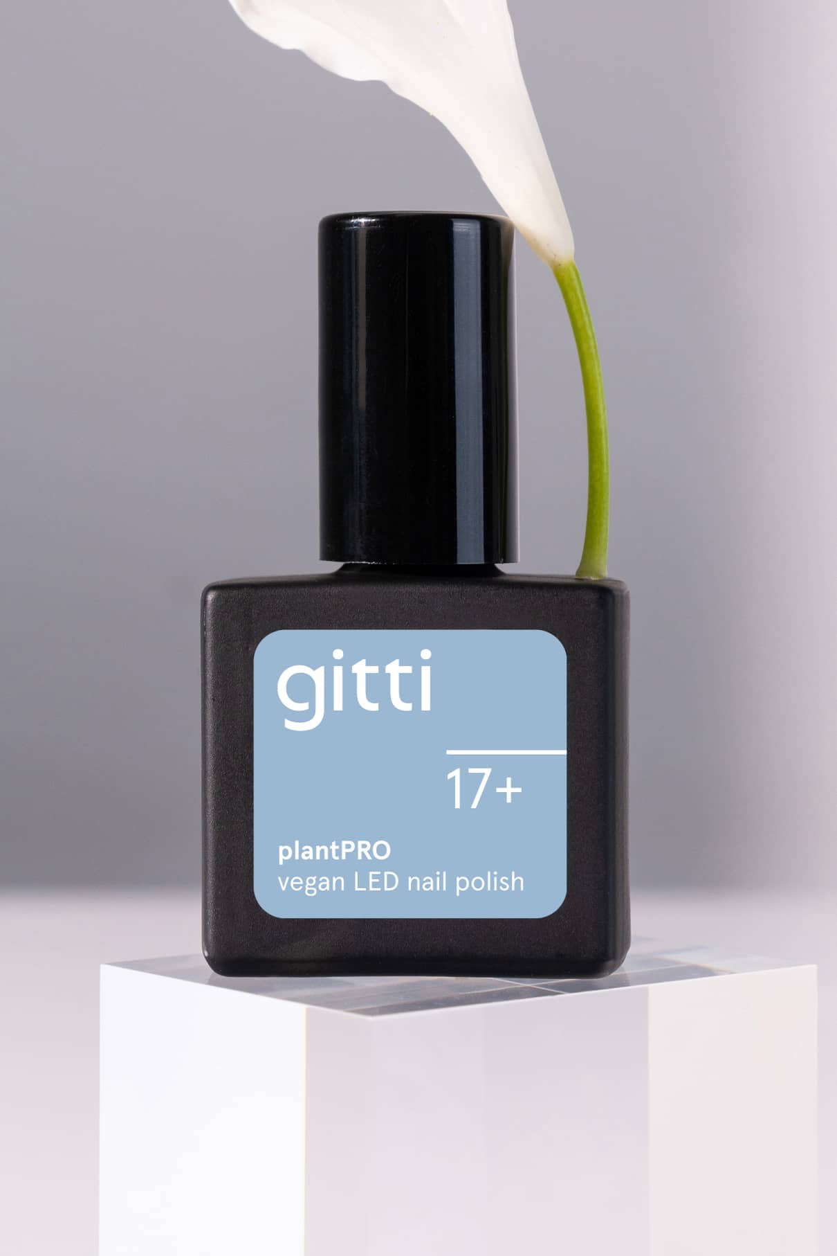 Ein schwarzes Fläschchen mit gitti no. 17+ plantPRO veganer LED-Nagellack mit einem Etikett in frischem Hellblau steht auf einem weißen Block, dahinter eine weiße Blume.