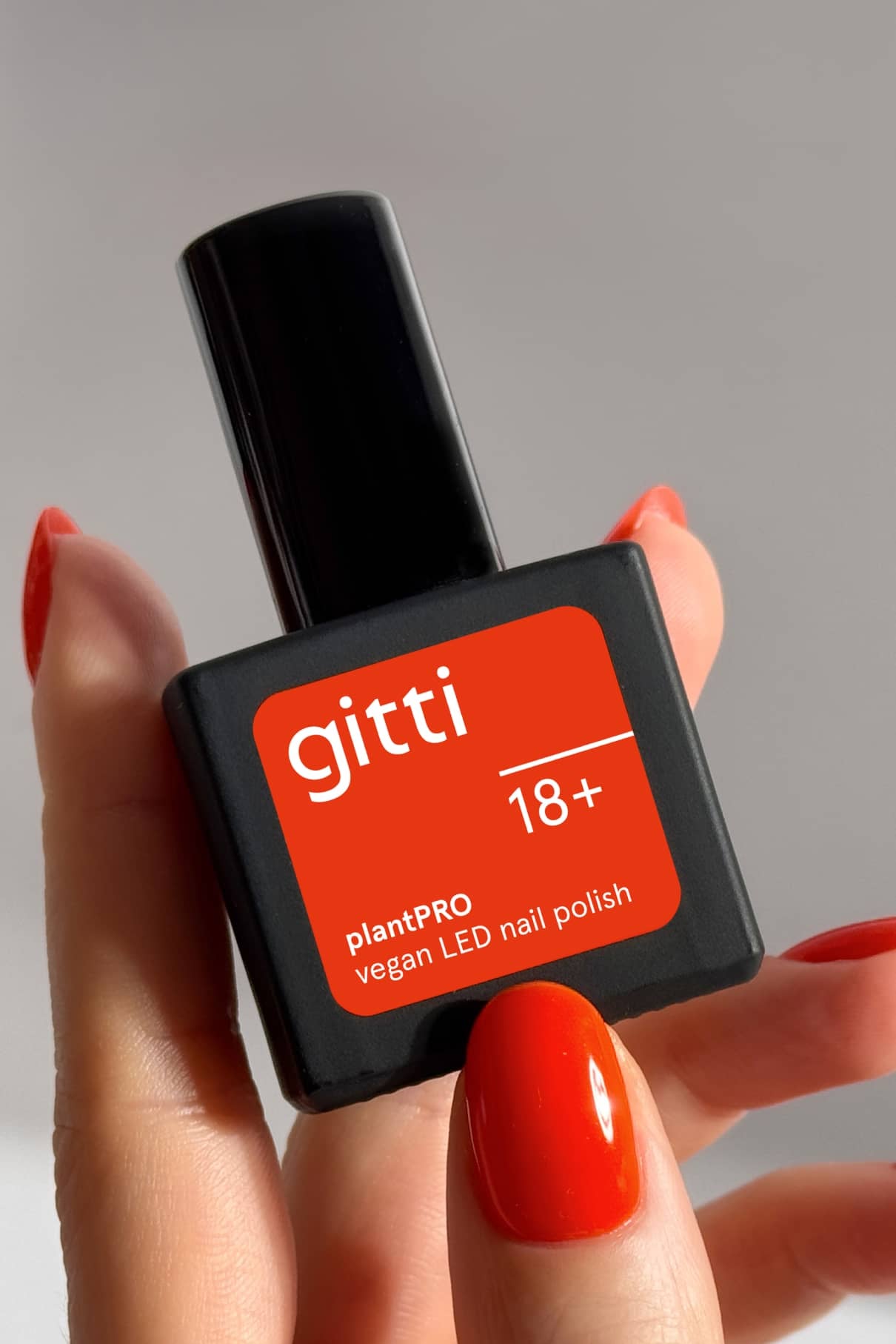Eine Hand mit rot lackierten Nägeln in leuchtendem Orange-Rot hält eine schwarze Flasche mit der Aufschrift gitti no. 18+ plantPRO vegan LED Nagellack.