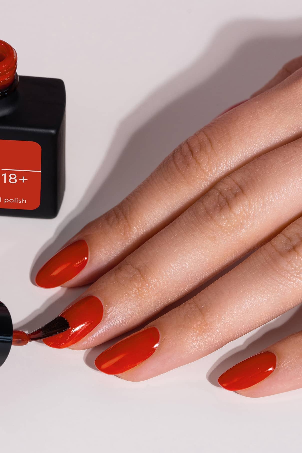 Eine Hand wird mit gitti no. 18+, einem leuchtenden, intensiv orange-roten Nagellack, während eine offene Flasche desselben Farbtons in der Nähe steht.