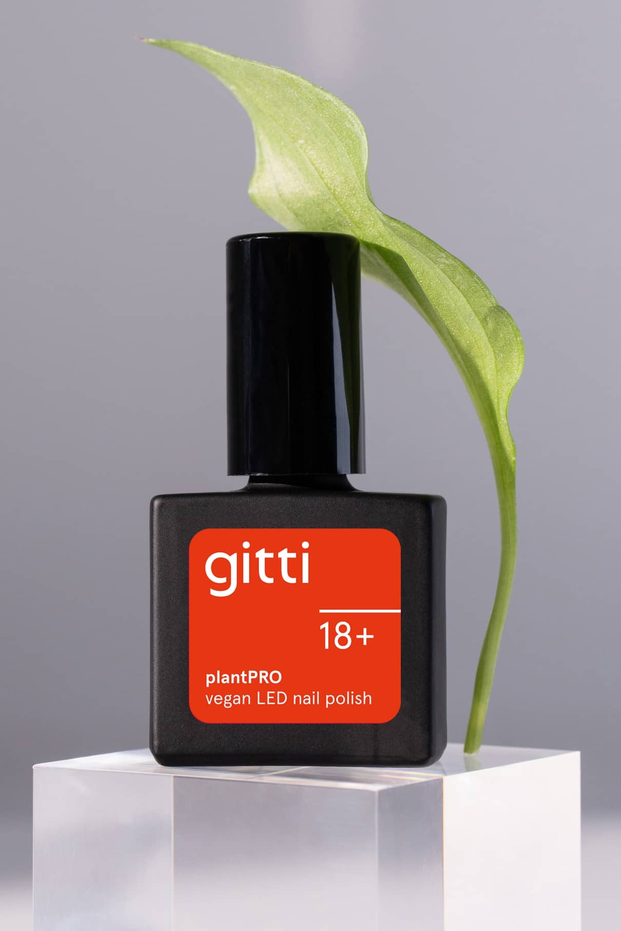 Eine Flasche des gitti no. 18+ plantPRO veganer LED-Nagellack in intensivem Orange-Rot steht auf einem durchsichtigen rechteckigen Block, dahinter ein grünes Blatt.