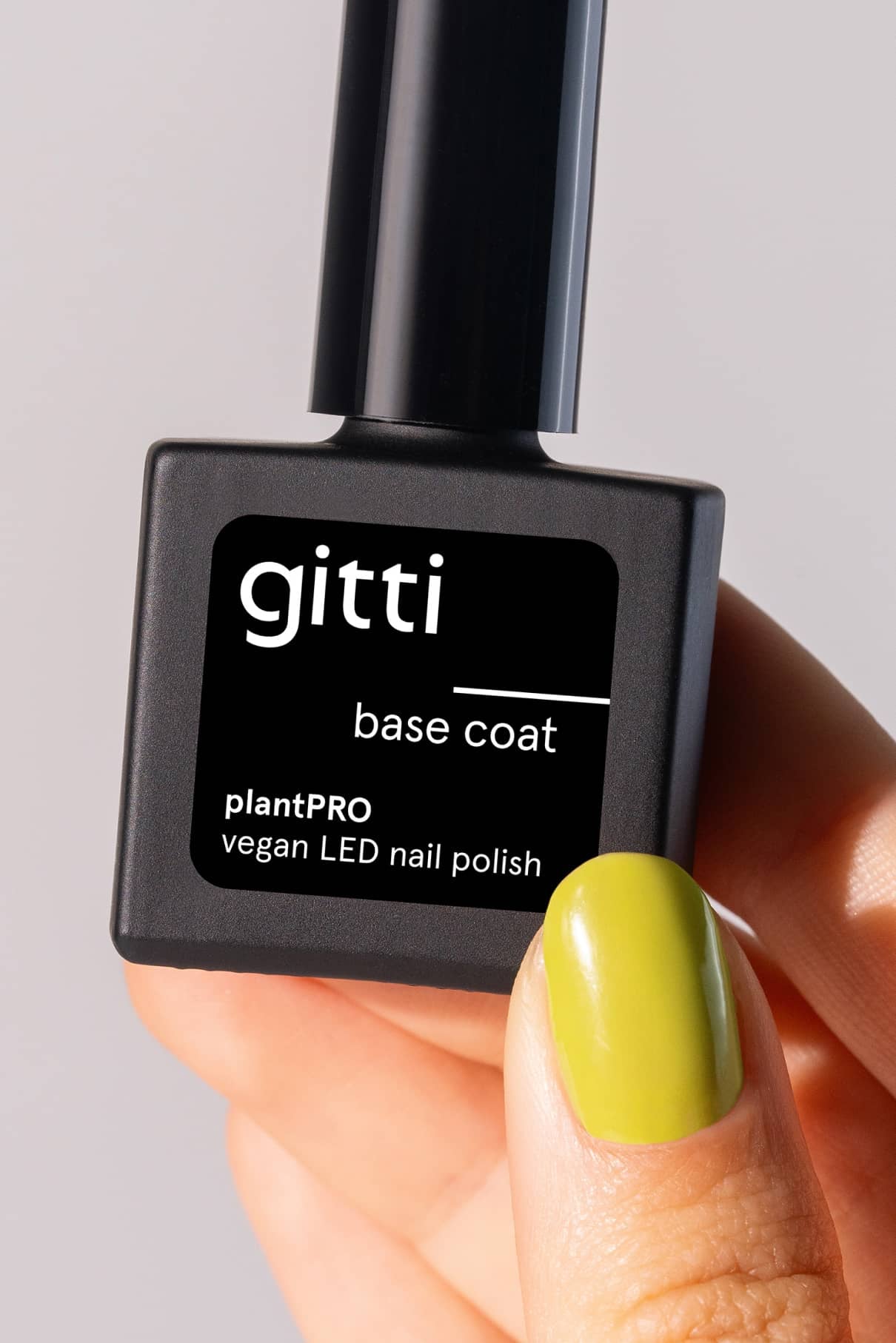 Eine Hand mit lindgrünem Nagellack hält eine Flasche plantPRO Base Coat, einen veganen LED-Gel-Nagelbasislack auf Pflanzenbasis, vor einem neutralen Hintergrund.