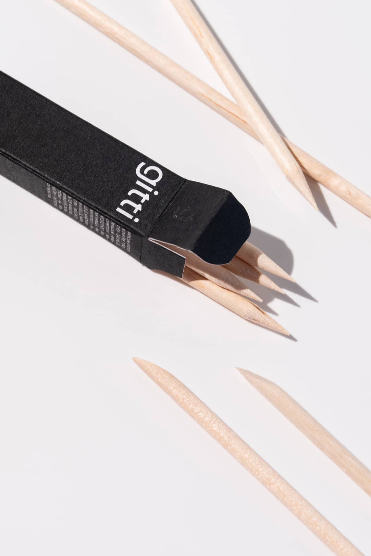Eine schwarze Schachtel mit der Aufschrift gitti PROsticks enthält unbehandelte Maniküre Sticks aus Birkenholz, wobei sich einige Sticks teilweise in der Schachtel befinden und einige daneben auf einer weißen Fläche liegen.