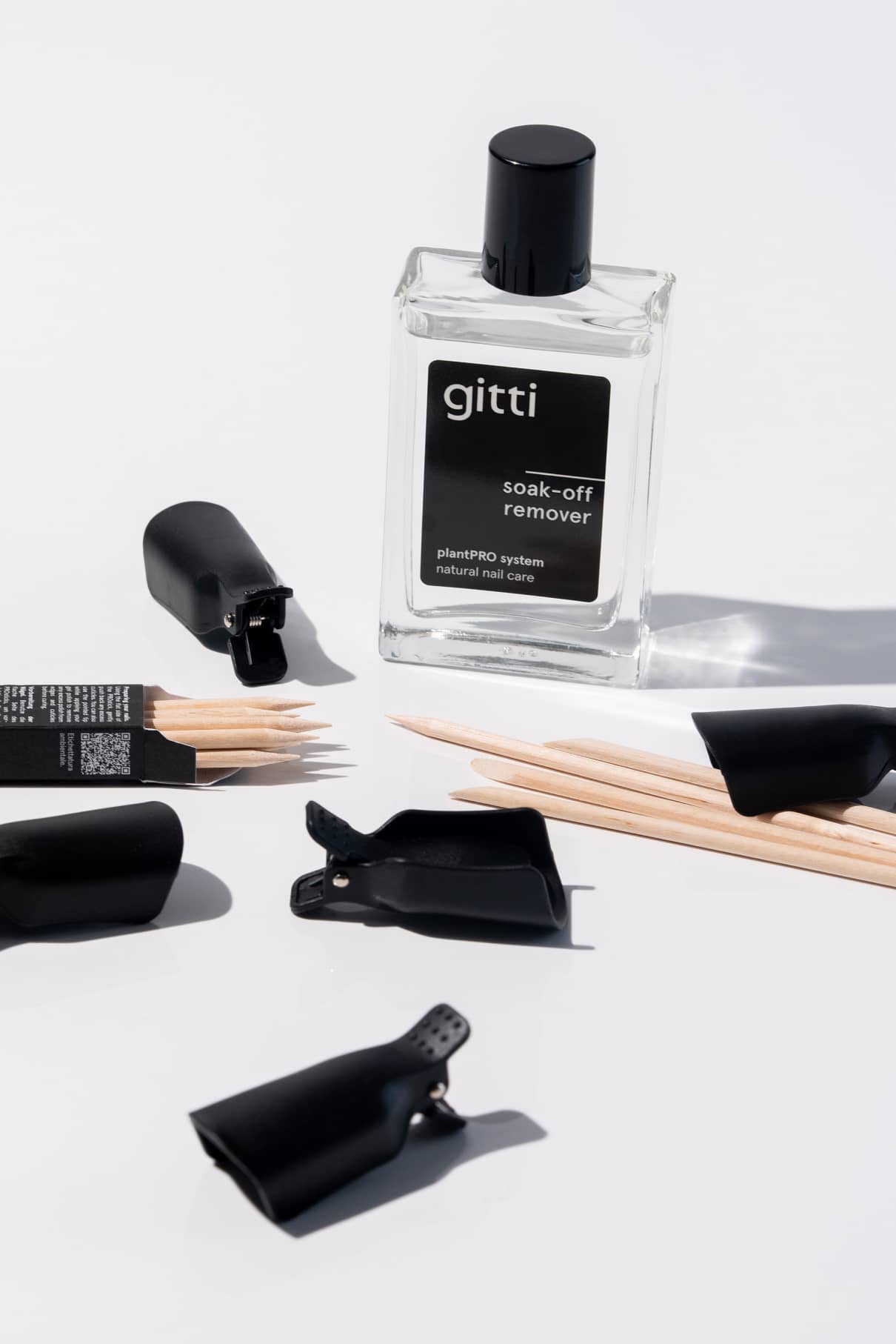 Eine Flasche Gitti Soak-Off-Entferner, PROsticks Maniküre Sticks aus Birkenholz und schwarze Nagelklammerhüllen sind auf einer weißen Fläche angeordnet.