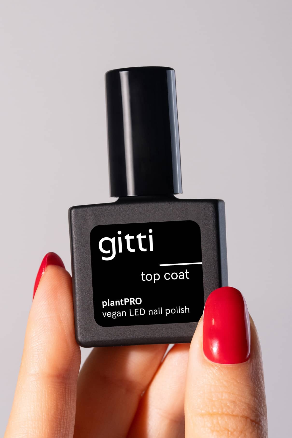 Eine Hand mit rot lackierten Nägeln hält eine schwarze Flasche plantPRO Top Coat, einen veganen Gellack auf Pflanzenbasis, vor einem schlichten Hintergrund.