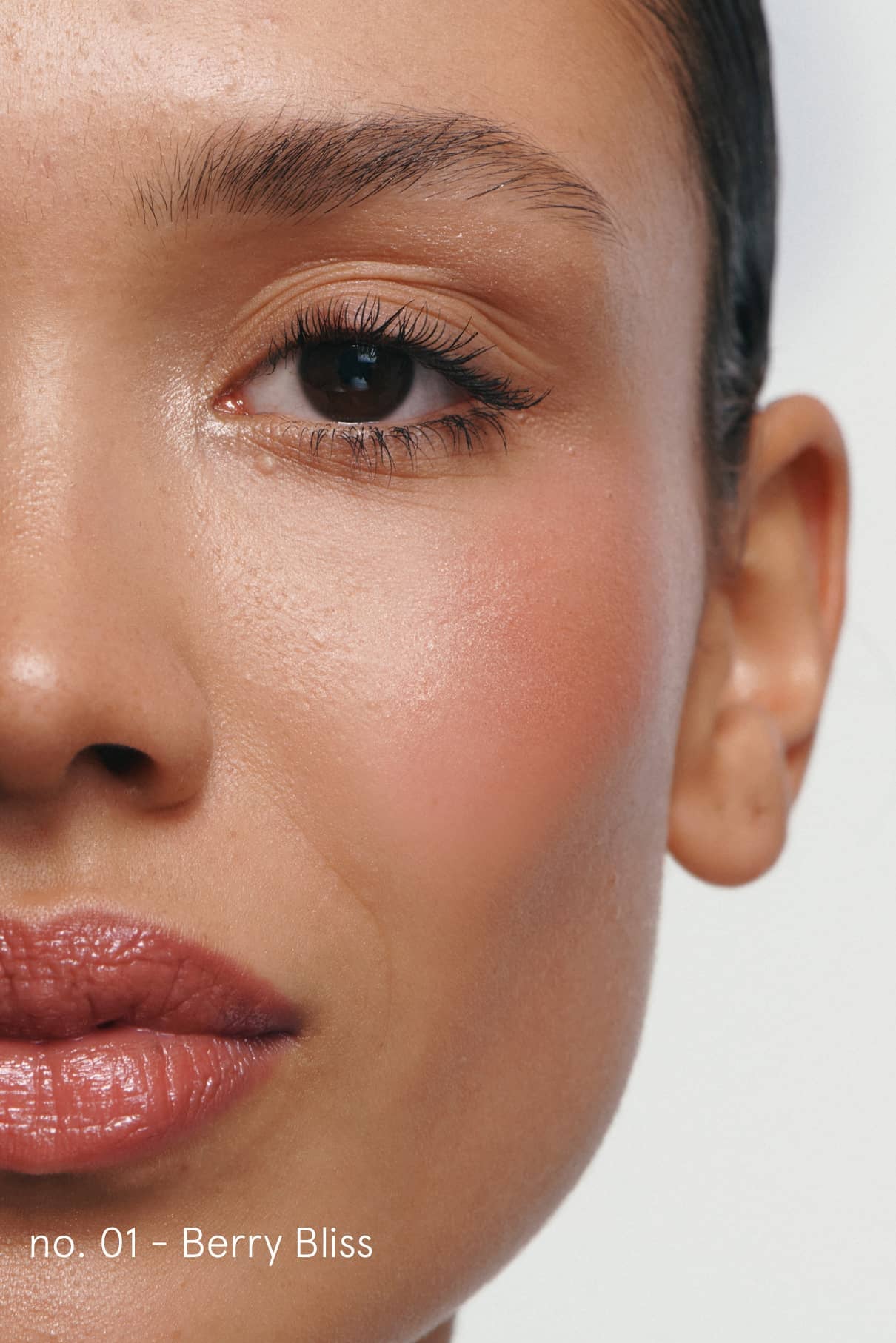 Nahaufnahme des Gesichts einer Frau mit glatter Haut, natürlichem Make-up, dezentem Rouge und glänzenden Lippen. Ihre Wimpern sind mit Cheek & Lash Duo Mascara geschminkt. Der Text am unteren Rand lautet Nr. 01 - Berry Bliss.
