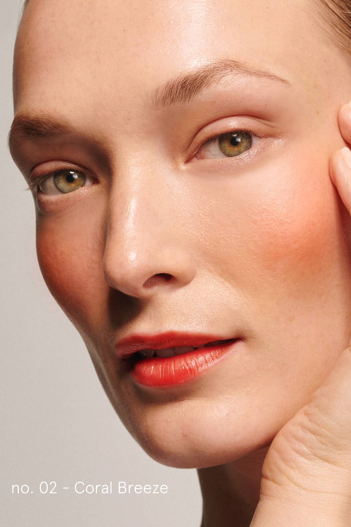 Eine Nahaufnahme einer Person mit heller Haut, die korallenrotes Rouge und Lippenstift trägt und den sanften Glanz von Cheek & Lash Duo in Nr. 02 - Coral Breeze zur Geltung bringt, wobei der Name dieser Nuance in der unteren linken Ecke erscheint.