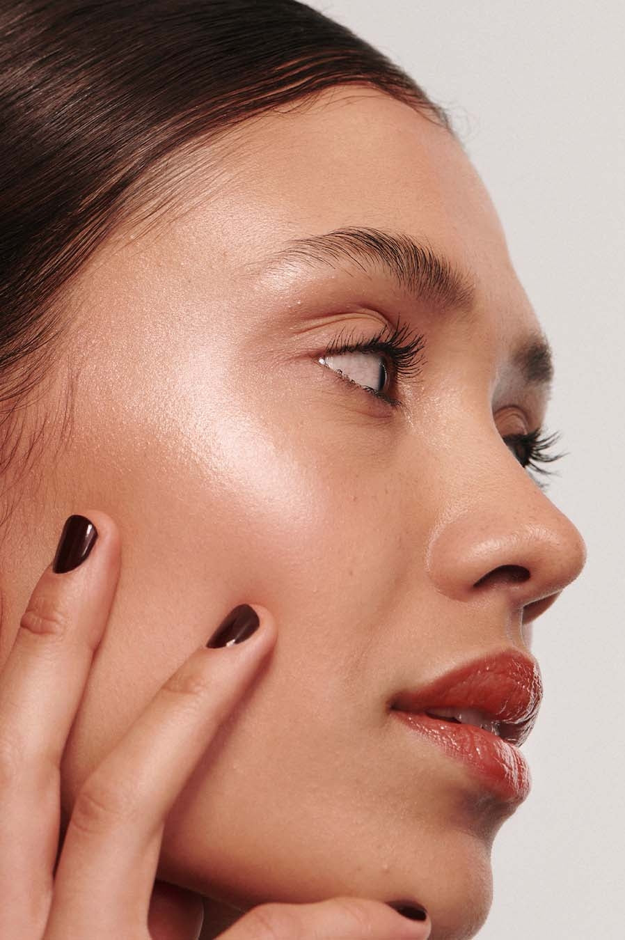 Nahaufnahme des Gesichts einer Person im Profil mit leuchtender, glatter Haut und mit Signature Set Lip Glaze verschönerten Lippen, wobei die dunklen Nägel sanft die samtenen Wangen berühren.