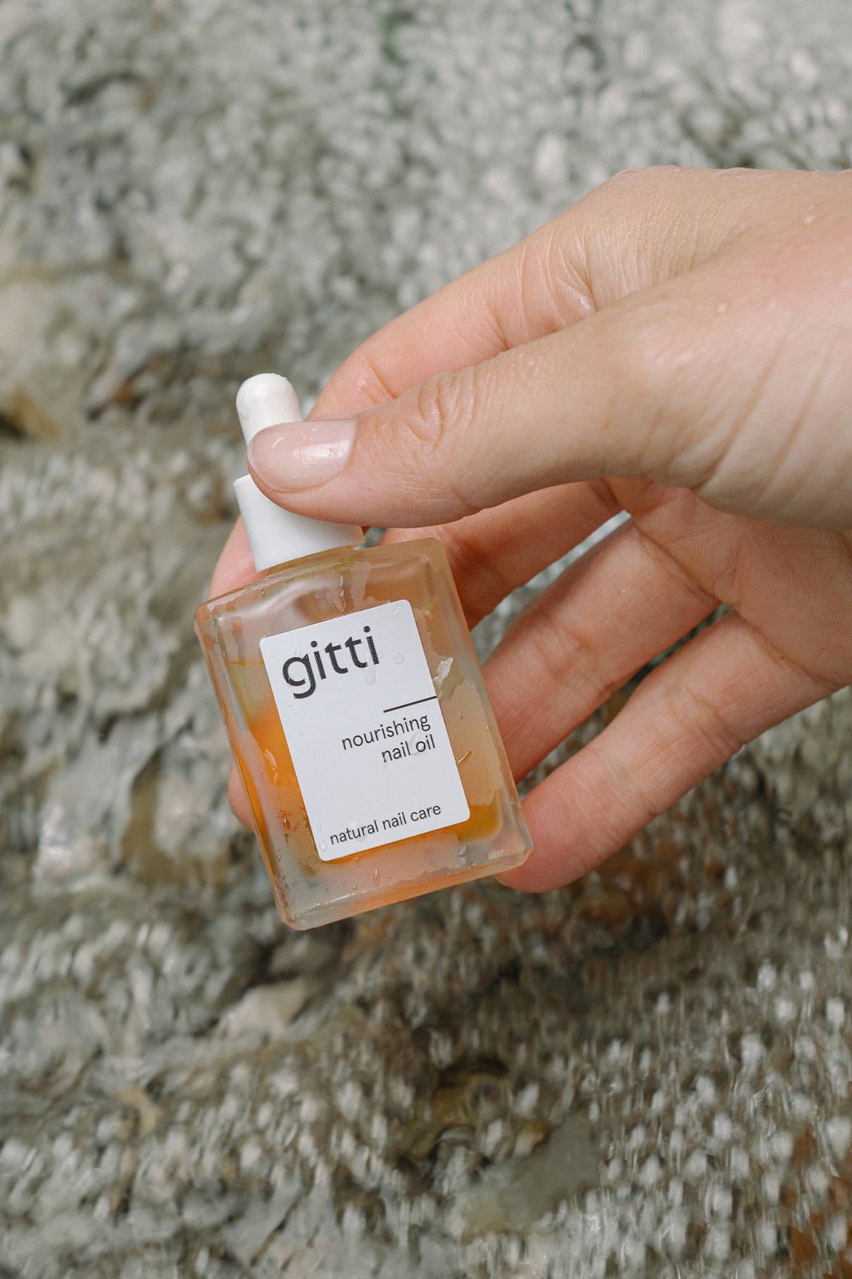 Eine Hand hält ein kleines Glasfläschchen aus dem Minimalist Manicure Set, das als pflegendes Nagelöl mit einem Tropfverschluss gekennzeichnet ist, vor einem strukturierten Hintergrund.