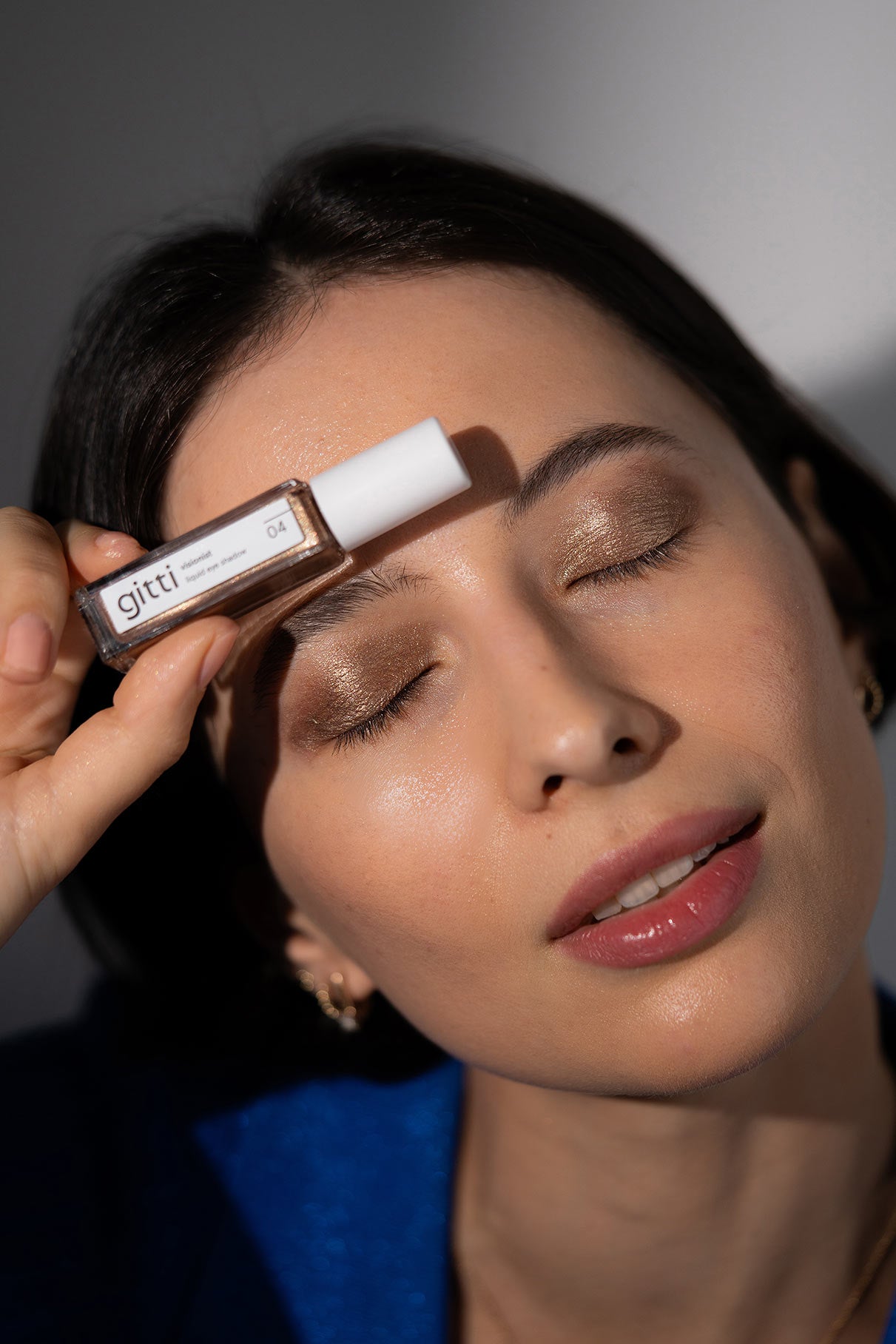 Eine Frau in einem blauen Oberteil mit schimmerndem Visionist Lidschatten-Augen-Make-up und voluminöser Wimperntusche hält eine Celebration Trio-Flasche mit Gitti-Nagelfarbe nahe ihrer Augenbraue, die Augen geschlossen.