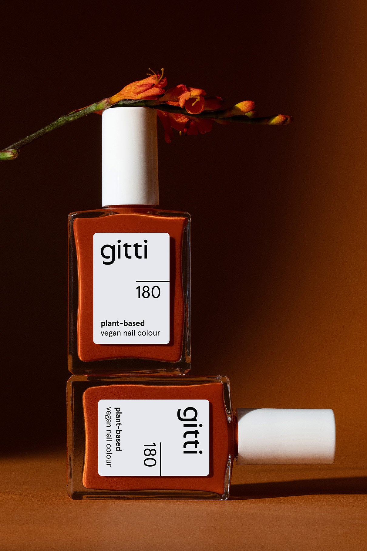Zwei Flaschen von gitti no. 180 pflanzlicher veganer Nagellack in einem satten Orange-Braun-Ton sind übereinander gestapelt, mit einer orangefarbenen Blume darüber und einem braunen Hintergrund.