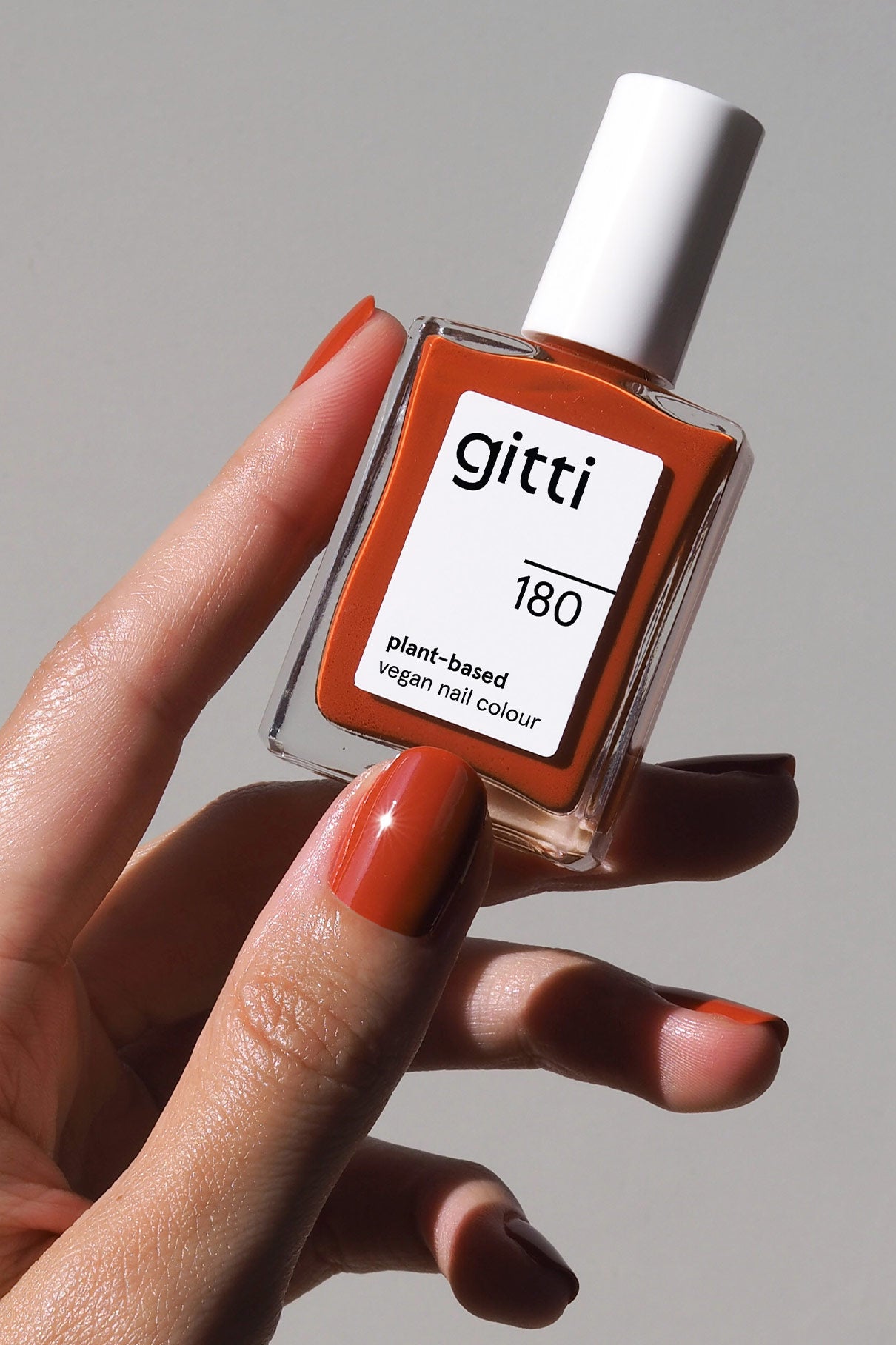 Eine Hand mit lackierten Nägeln hält ein Fläschchen mit gitti no. 180 pflanzlicher veganer Nagelfarbe in leuchtendem Orange-Braun vor einem schlichten Hintergrund.