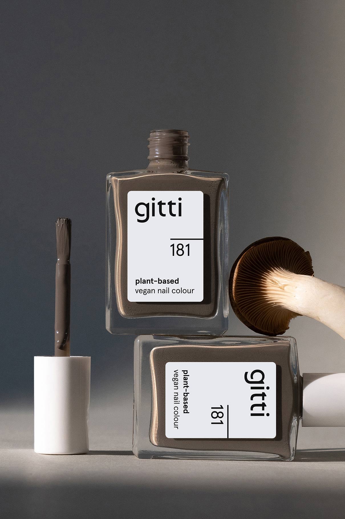 Zwei Flaschen von gitti no. 181 pflanzliche, vegane Nagelfarbe mit einem schönen graubraunen Unterton - eine ist offen, der Pinsel liegt daneben, und neben den taupefarbenen Flaschen steht ein Pilz.