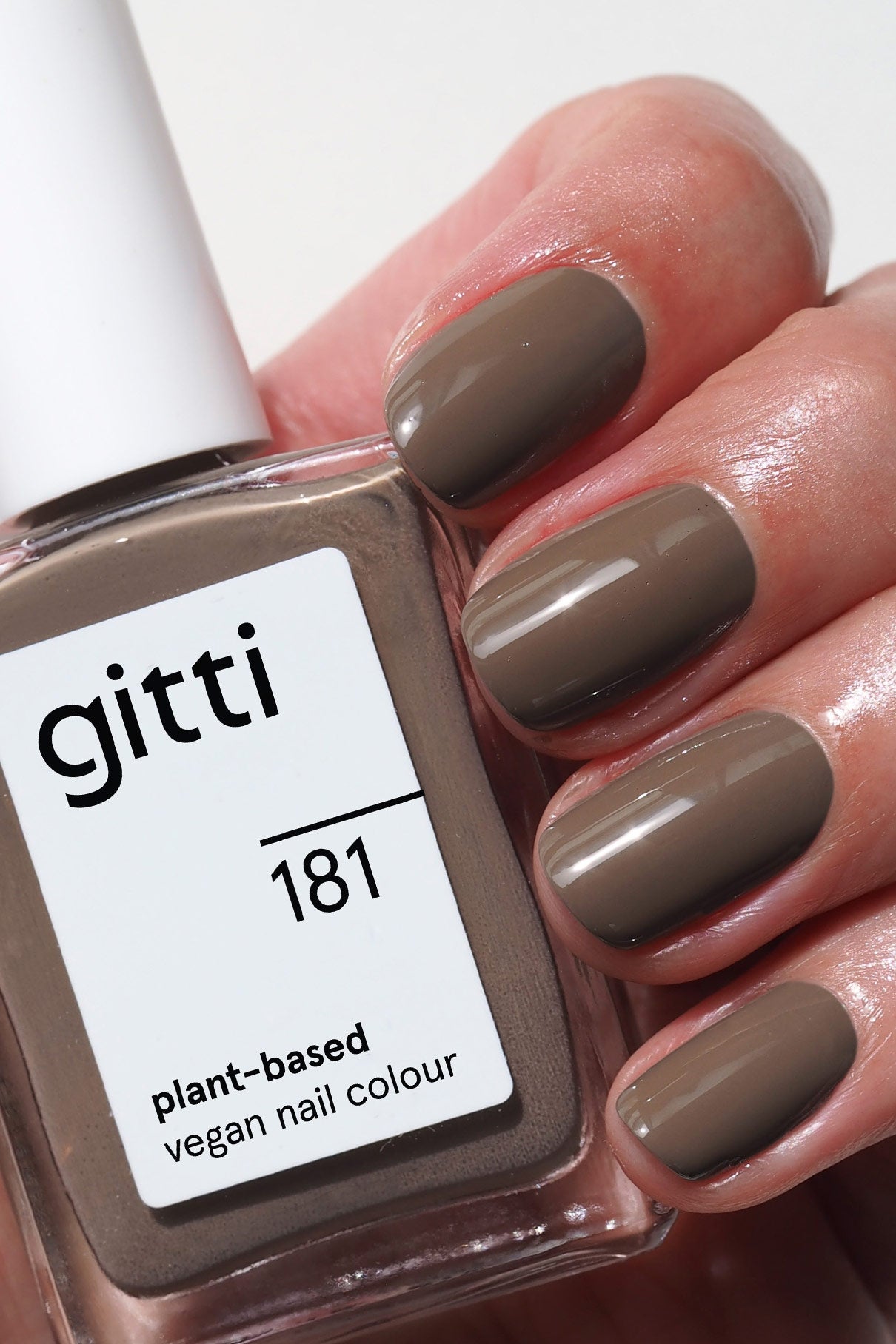 Eine Hand mit kurzen, in kühlem Graubraun-Taupe lackierten Nägeln hält eine Flasche gitti no. 181, einem pflanzlichen, veganen Nagellack im passenden Farbton.