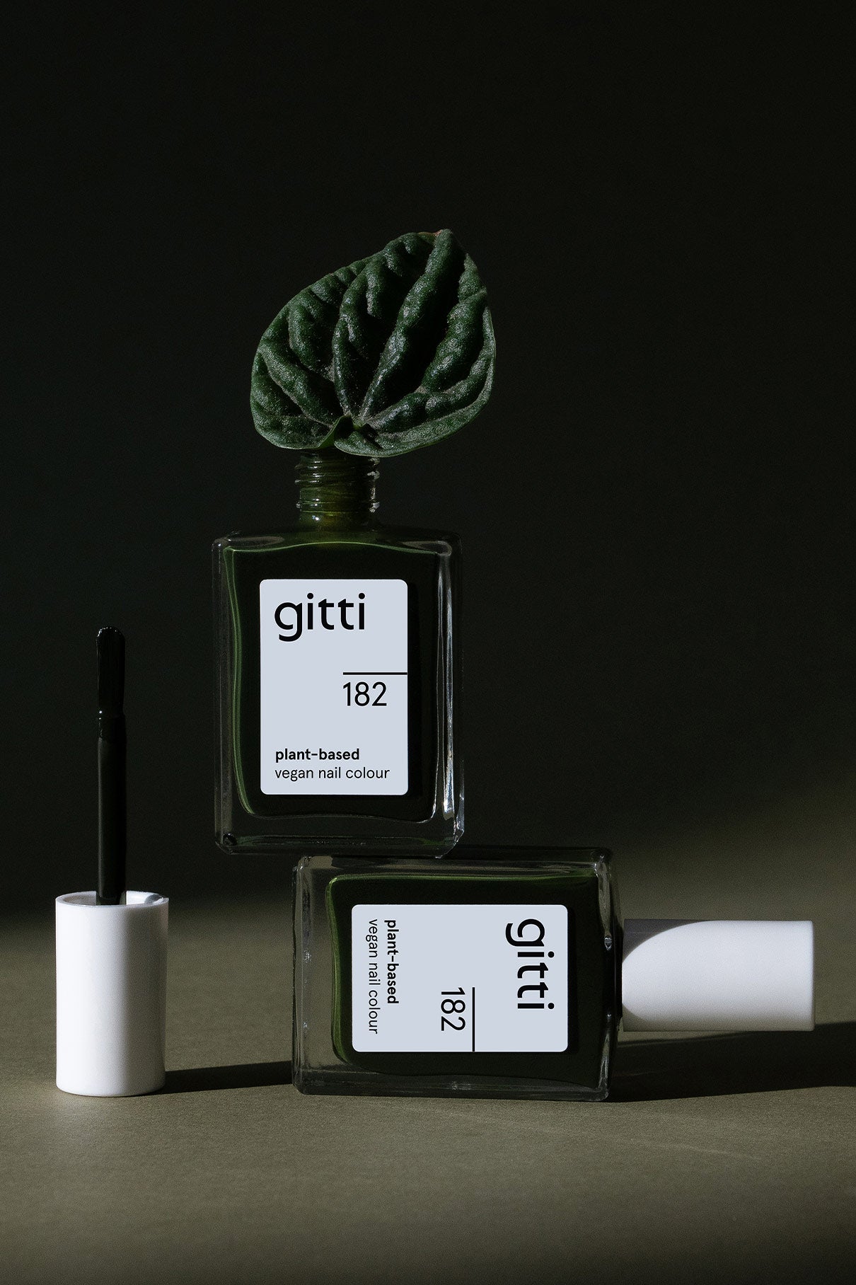 Zwei Flaschen von gitti no. 182 pflanzlicher, veganer Nagellack - eine aufrecht mit einem Blatt darauf, die andere liegend neben einem offenen Pinsel in dramatischem Licht - zeigt den tiefgrünen Farbton.