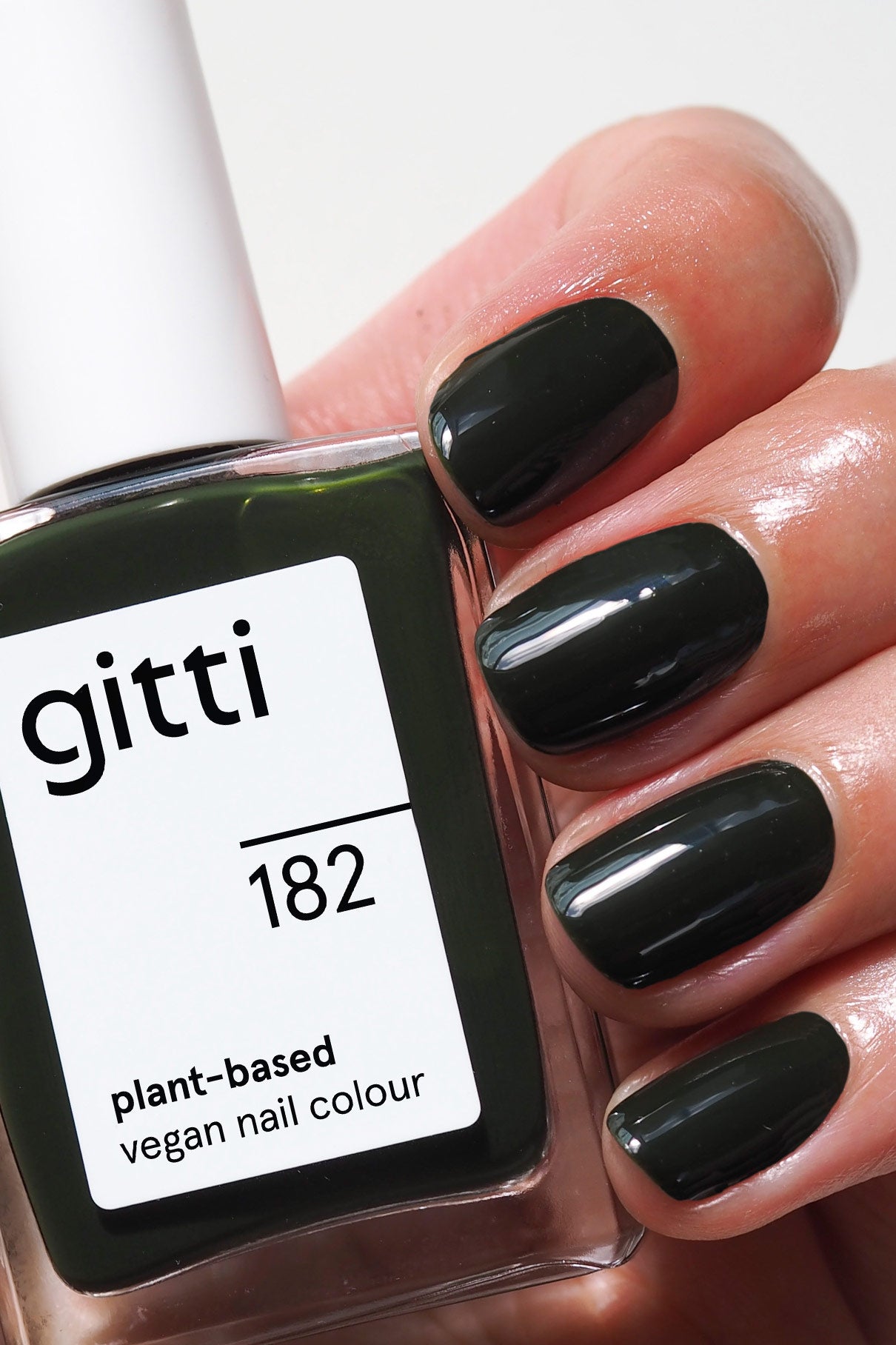 Eine Hand mit kurzen, sauber manikürten und in dunklem Tannengrün lackierten Nägeln hält eine Flasche mit der Aufschrift gitti no. 182, vegane Nagelfarbe auf Pflanzenbasis.