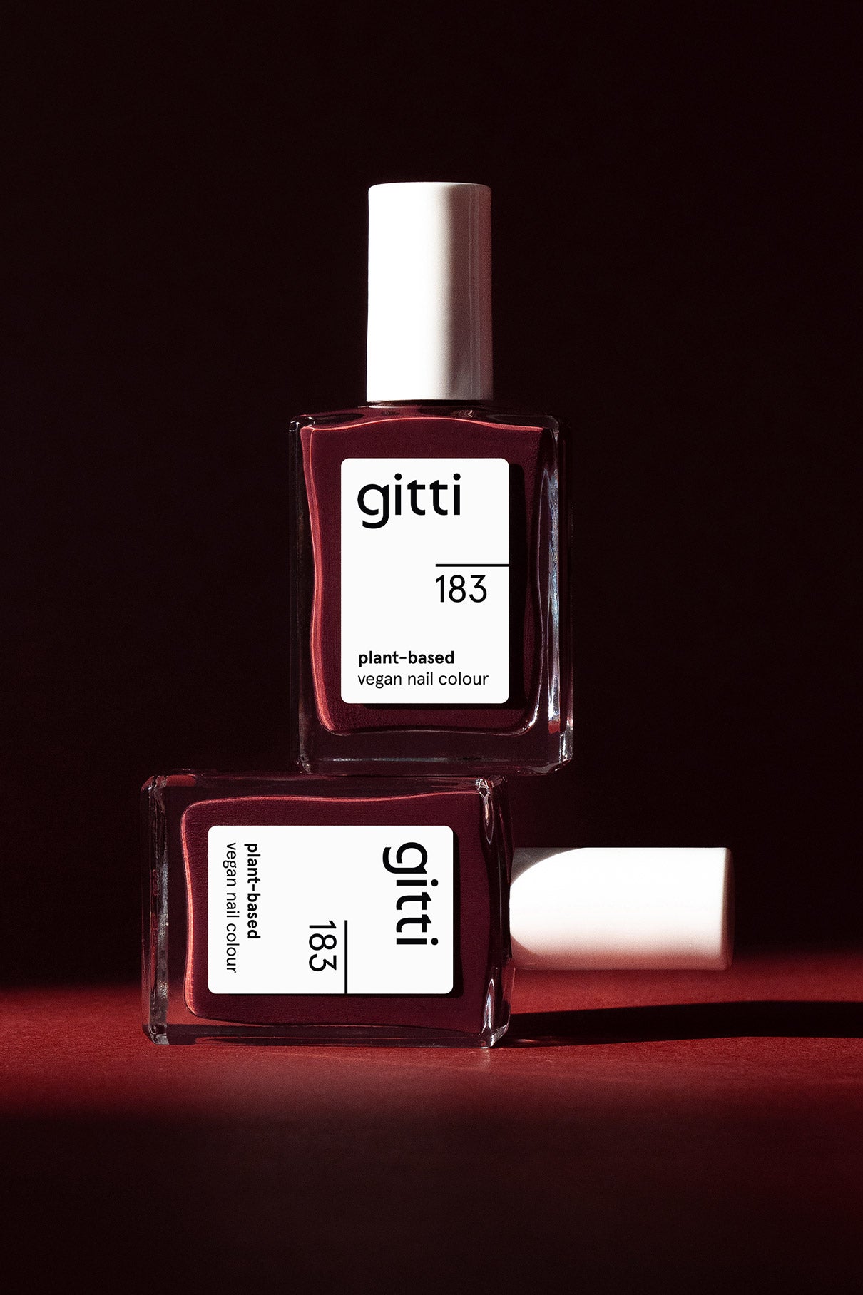 Zwei Flaschen gitti no. 183 pflanzliche vegane Nagelfarbe in intensivem Beerenrot mit weißen Kappen - eine stehend, eine liegend - stehen vor einem dunkelroten Hintergrund.