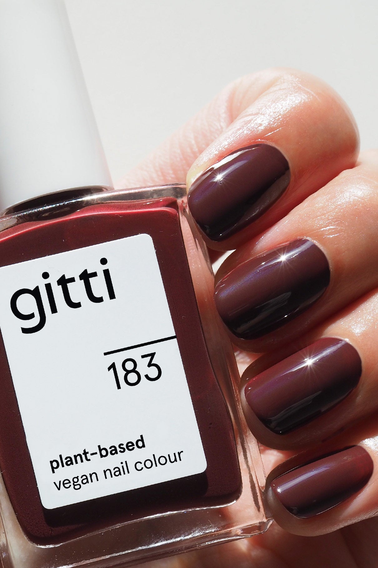 Eine Hand mit sauber manikürten, in dunklem Beerenrot lackierten Nägeln hält eine Flasche gitti no. 183, einer veganen Nagelfarbe auf Pflanzenbasis.