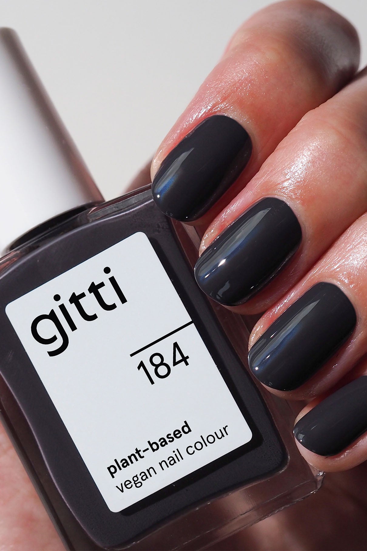 Eine Hand mit kurzen, quadratisch geformten Nägeln, die in einem glänzenden dunklen Anthrazit lackiert sind, hält eine Flasche mit der Aufschrift gitti no. 184, vegane Nagelfarbe auf Pflanzenbasis.