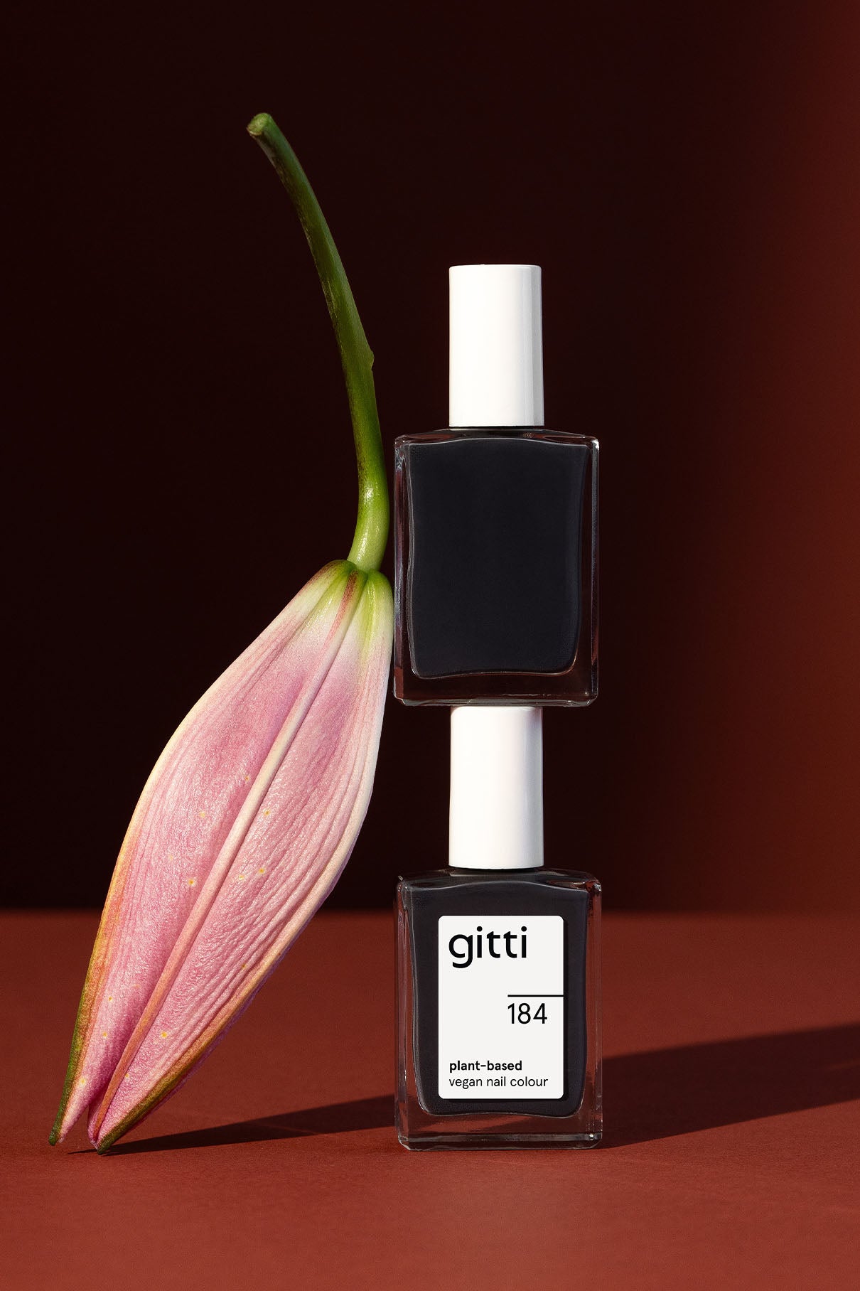Zwei quadratische Flaschen von gitti no. 184 Nagellack in dunklem Anthrazit sind neben einer rosa Lilienknospe vor dunkelrotem Hintergrund gestapelt.
