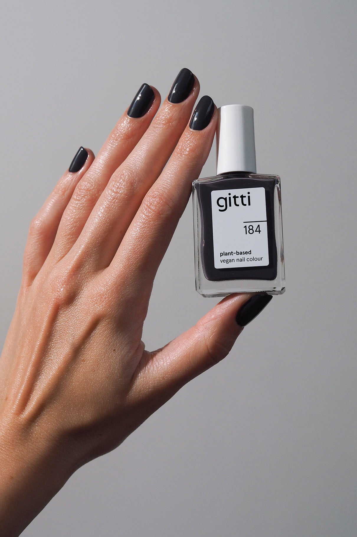 Eine Hand mit dunkel gefärbten Nägeln hält ein Fläschchen mit gitti no. 184, einem pflanzlichen, veganen Nagellack in dunklem Anthrazit, vor einem schlichten Hintergrund.