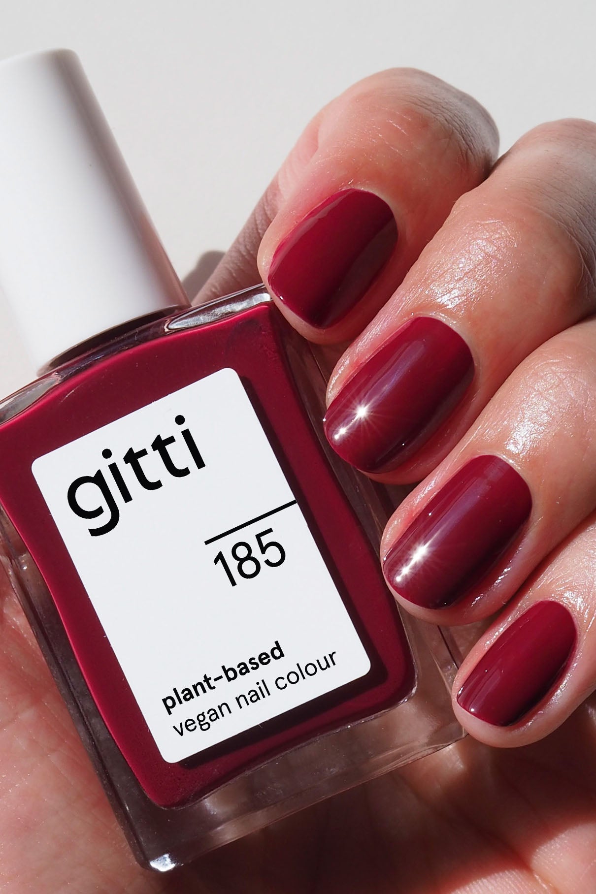 Eine Hand mit lackierten Nägeln hält eine Flasche von gitti no. 185, einem veganen Nagellack auf Pflanzenbasis in tiefem Rot mit kühlen Untertönen.