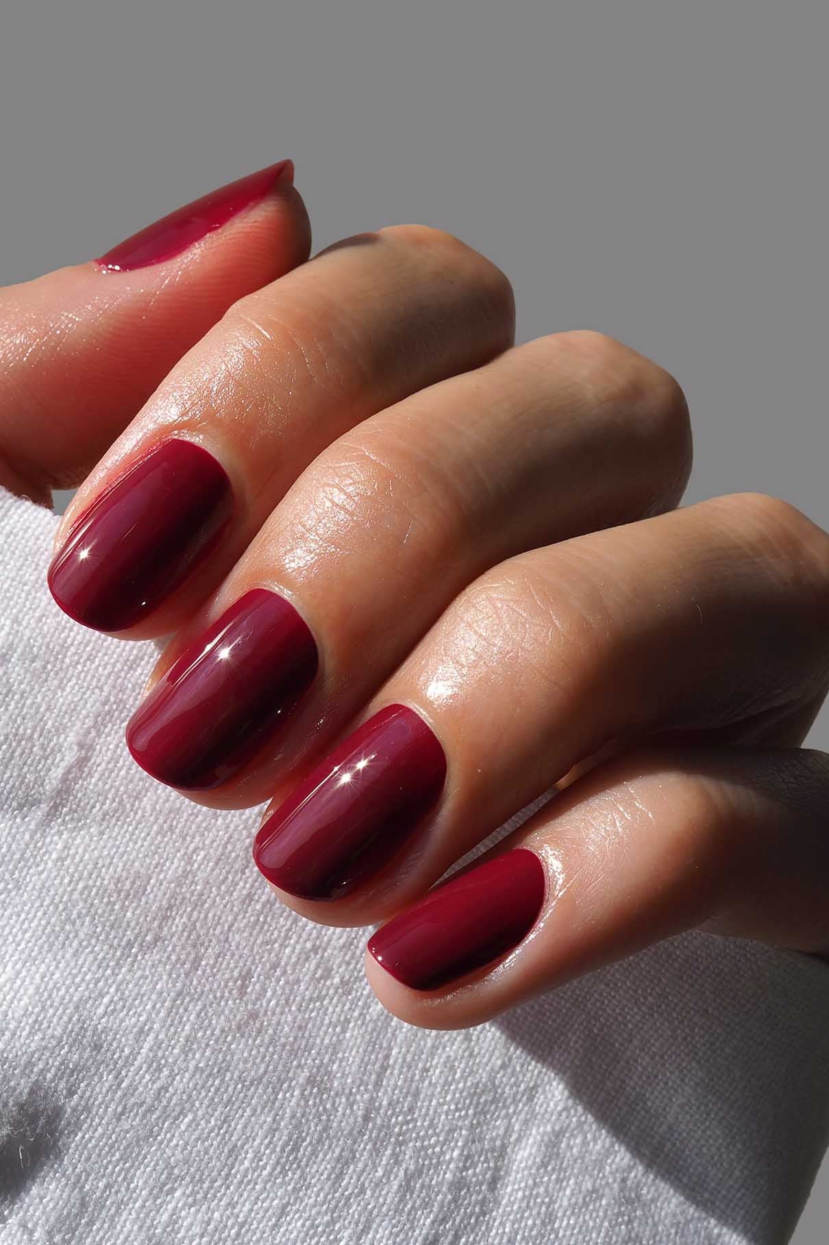 Eine Hand mit sauber manikürten Nägeln, die in dem glänzenden, kühl getönten, tiefen Rot von gitti no. 185 lackierten Nägeln hält einen weißen Stoff vor einem schlichten grauen Hintergrund.
