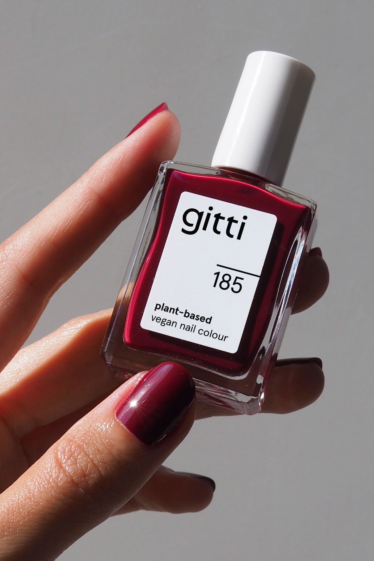 Eine Hand mit lackierten Nägeln hält eine Flasche der pflanzlichen veganen Nagelfarbe Nails + Lips in Sync in einem tiefroten Farbton vor einem neutralen Hintergrund.