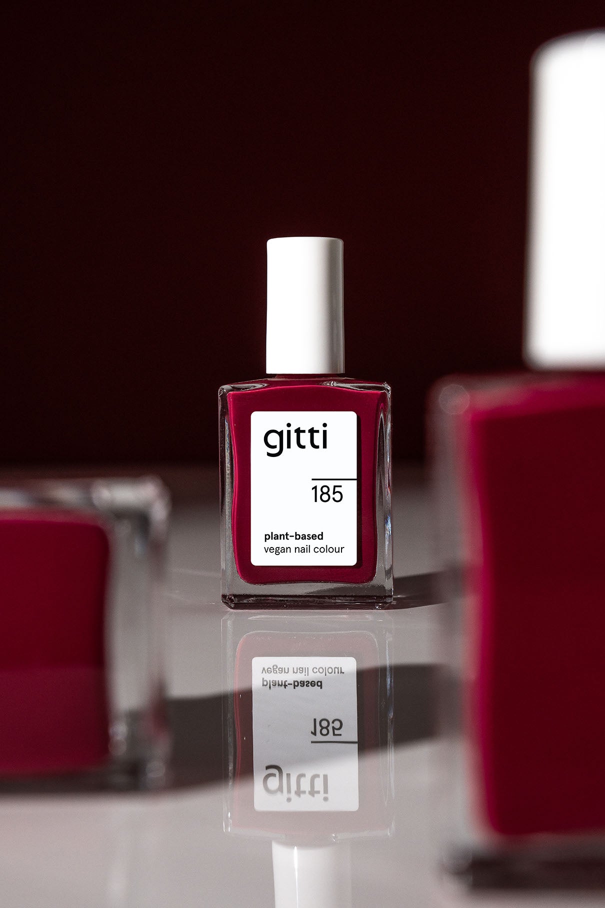 Eine Flasche von gitti no. 185, eine vegane Nagelfarbe auf pflanzlicher Basis in tiefem Rot mit kühlen Untertönen, steht auf einer spiegelnden Oberfläche mit verschwommenen Flaschen im Vorder- und Hintergrund.