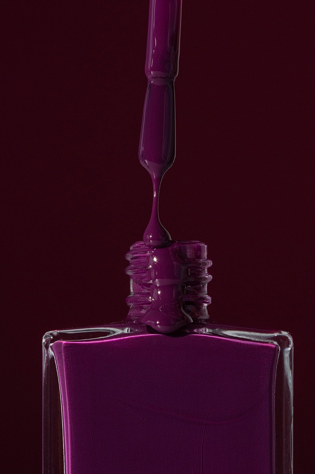 Eine Nahaufnahme eines Pinsels, der lila Gitti No. 186 Nagellack in eine viereckige Flasche tropft, vor einem dunkelvioletten und smaragdgrünen Hintergrund.