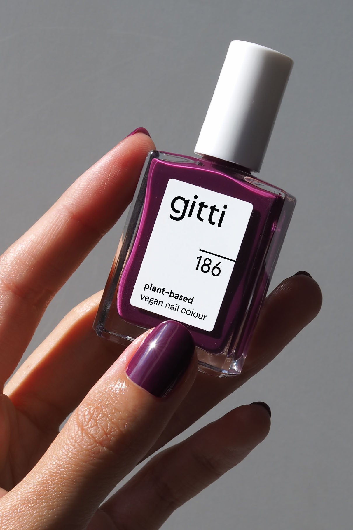 Eine Hand mit lila lackierten Nägeln hält eine Flasche gitti no. 186 pflanzliche vegane Nagelfarbe in Smaragdgrün vor einem neutralen Hintergrund.