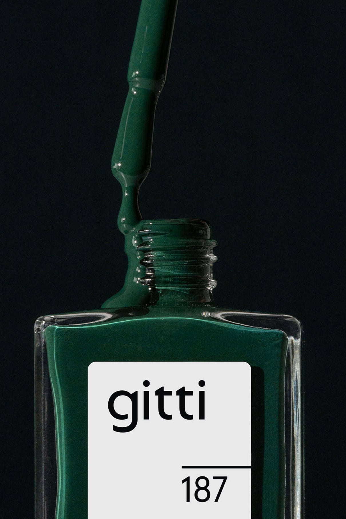 Ein grüner Nagellackpinsel schwebt über einer offenen rechteckigen Glasflasche mit der Aufschrift gitti no. 187, wobei intensiver Smaragdgrüner Nagellack in die Flasche zurücktropft.