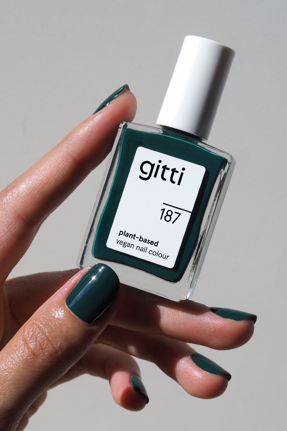 Eine Hand mit dunkelgrün lackierten Nägeln in gitti no. 187 hält eine Flasche mit gitti no. 187 pflanzlicher veganer Nagelfarbe vor einem neutralen Hintergrund.