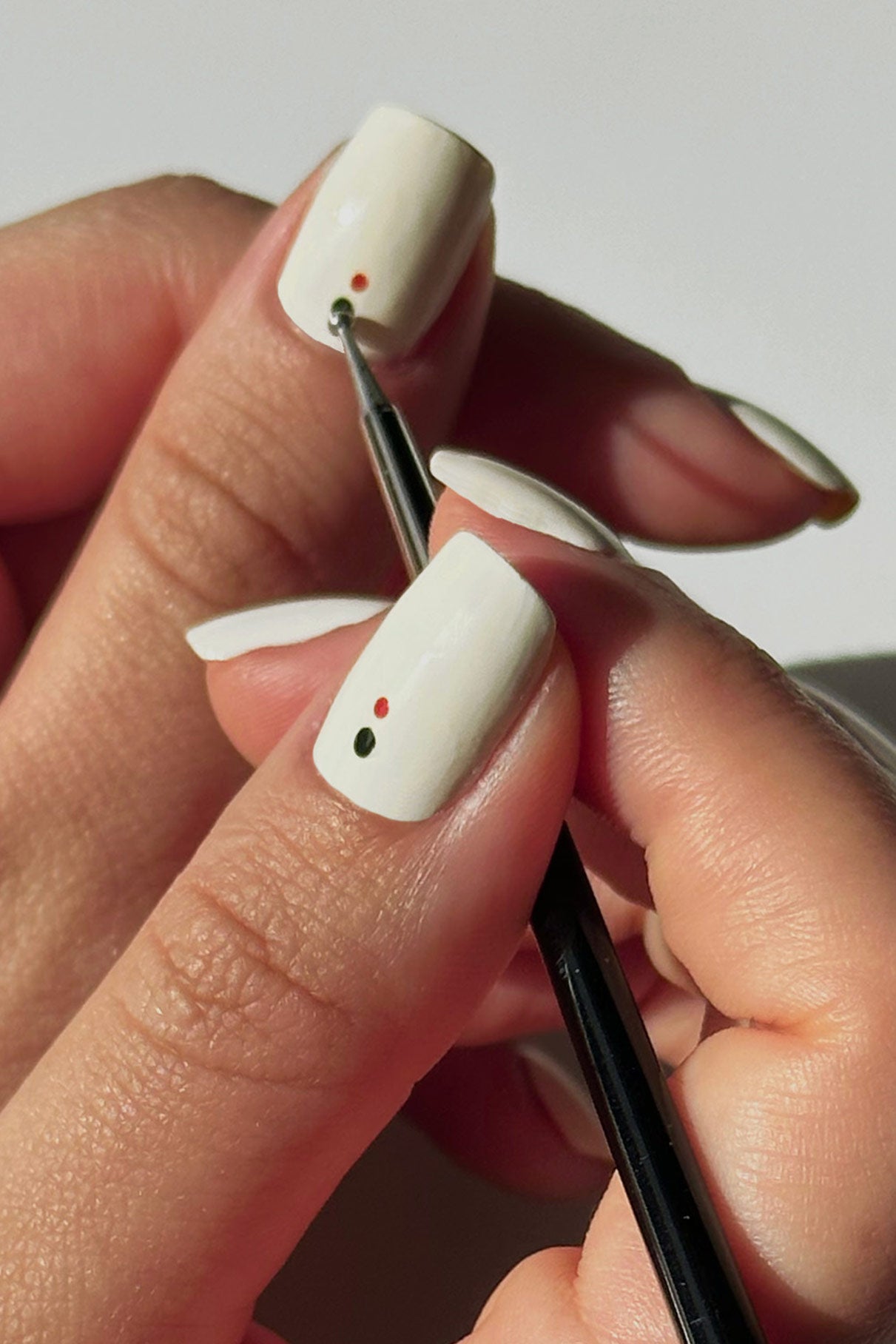 Eine Hand mit weiß lackierten Nägeln wird mit zwei kleinen Punkten, einem roten und einem grünen, verziert, wobei der präzise Nail Art Dotter für kreative Nail Art verwendet wird.