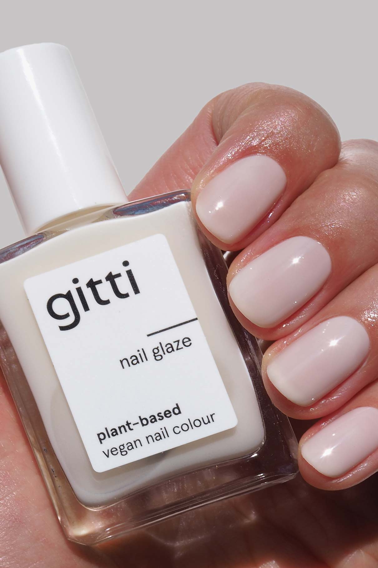 Eine Hand mit sauber manikürten, blassrosa Nails + Lips in Sync-Nägeln hält die passende pflanzliche vegane Nagellackierung vor einem hellgrauen Hintergrund.