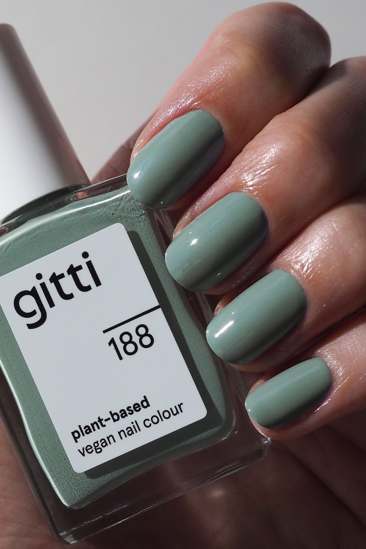 Eine Hand mit sauber manikürten, in einem gedeckten Grünton lackierten Nägeln hält eine Flasche veganen Nagellack auf Pflanzenbasis von Nails + Lips in Sync, der perfekt zur Farbe passt.