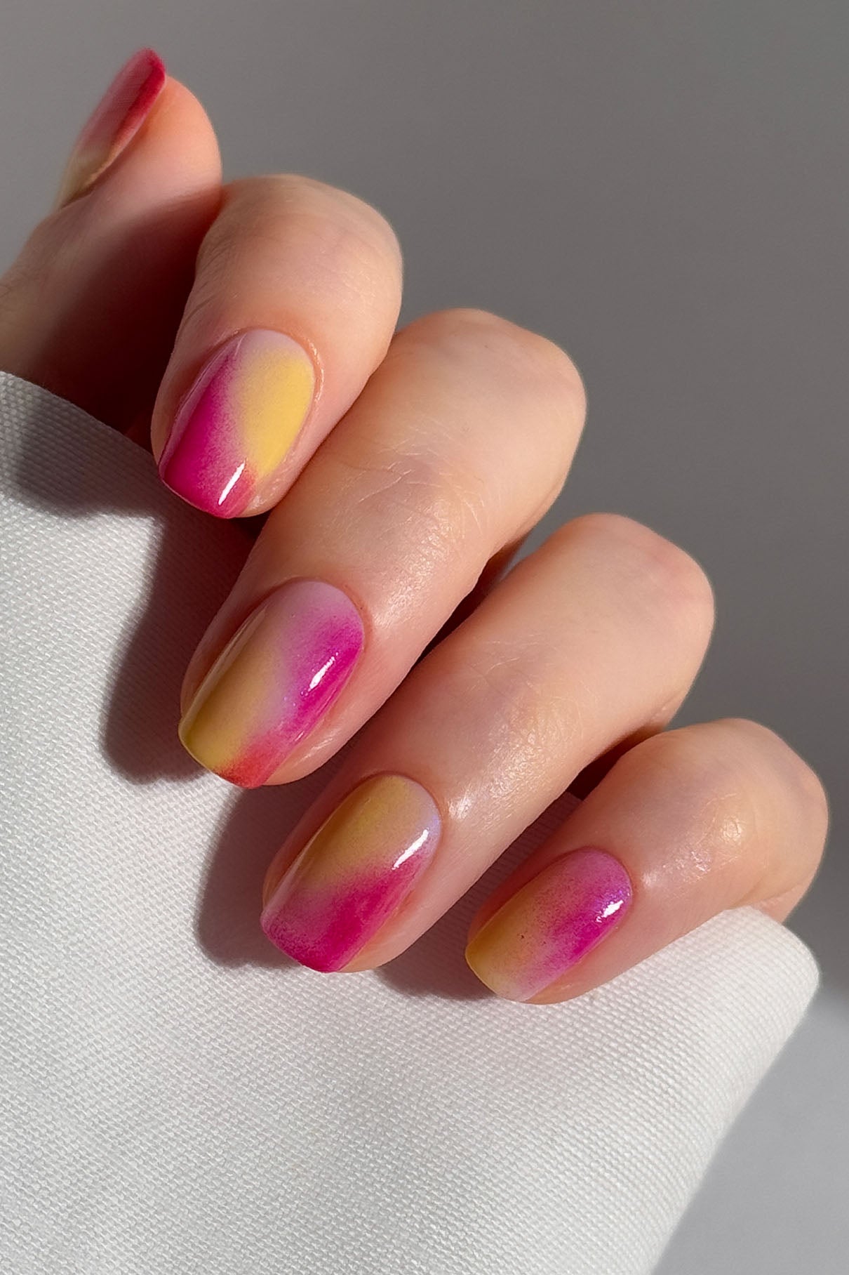 Eine Hand mit kurzen, ovalen Nägeln zeigt ein rosa und gelbes Ombre-Design, das mit dem Nailart-Set erstellt wurde, vor einem weißen Stoffhintergrund.