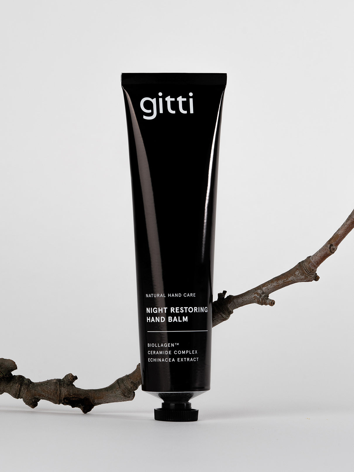 Eine schwarze Tube des Night Restoring Hand Balm steht aufrecht auf einem weißen Hintergrund, gestützt von einem trockenen Zweig - die ideale Pflege für Ihre Hände über Nacht.