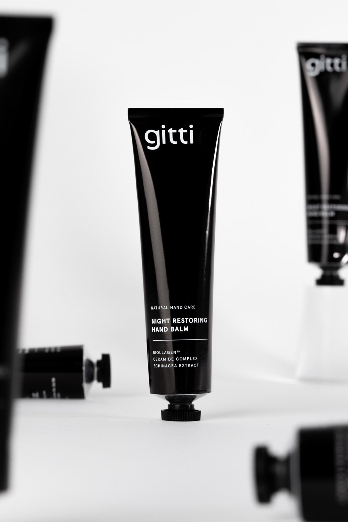 Eine schwarze Tube mit der Aufschrift Night Restoring Hand Balm steht aufrecht, umgeben von ähnlichen Tuben auf weißem Hintergrund - die ideale Handcreme für deine nächtliche Pflege und perfekte Regeneration deiner Hände über Nacht.