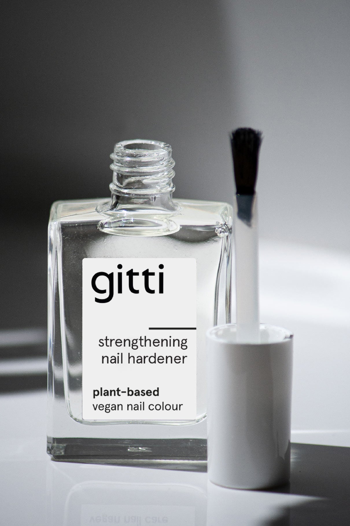 Eine durchsichtige Flasche des Strengthening Nail Hardener, einer pflanzlichen und veganen Nagelfarbe für stärkere Nägel, wird mit dem Pinsel auf einer weißen Fläche geöffnet gezeigt - ideal für brüchige Nägel.