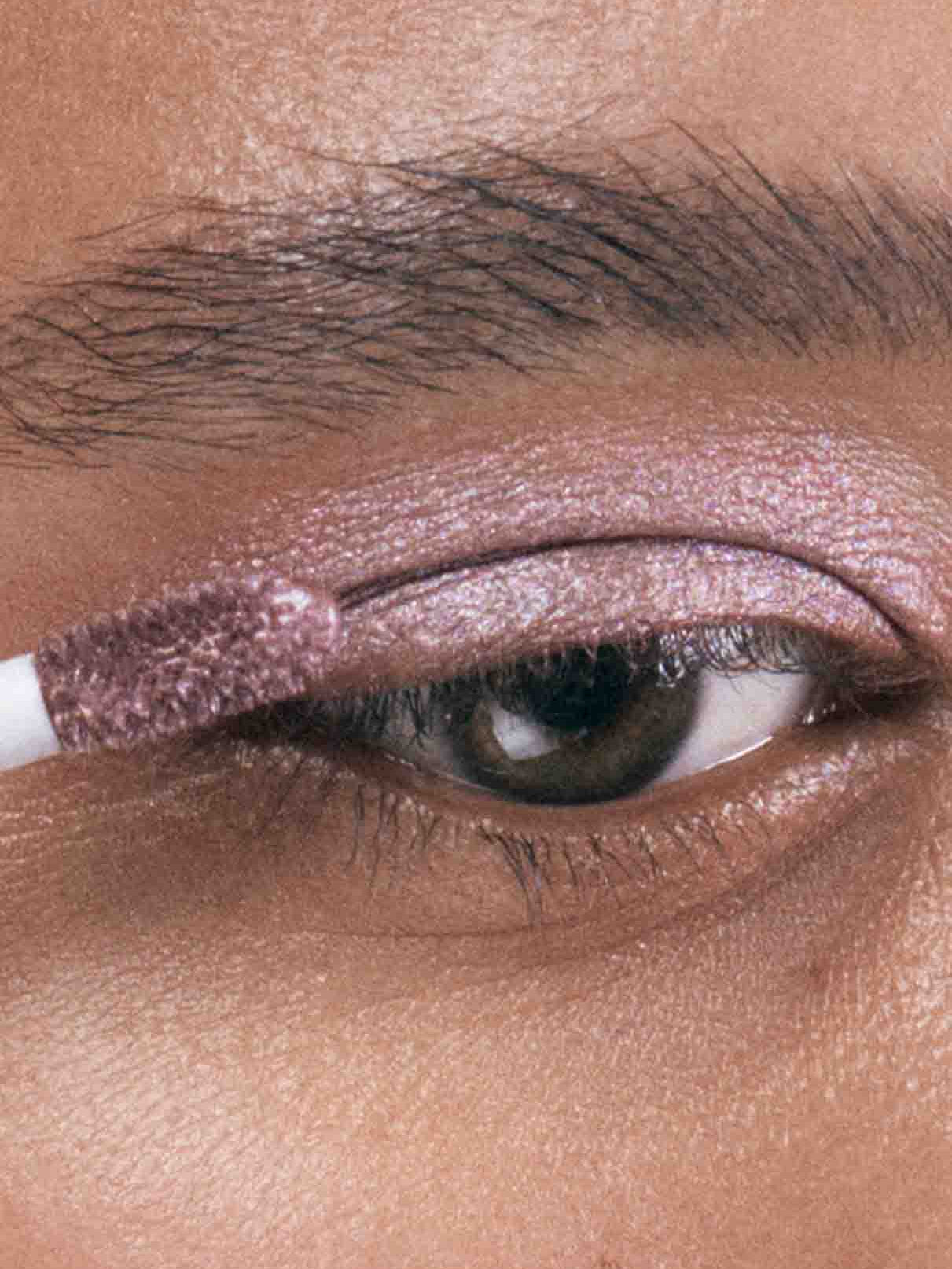 Eine Nahaufnahme eines Auges, während Visionist Nr. 01, ein schimmernder, beerenfarbener Lidschatten mit kühlem Unterton, mit einem Make-up-Applikator auf das Augenlid aufgetragen wird.