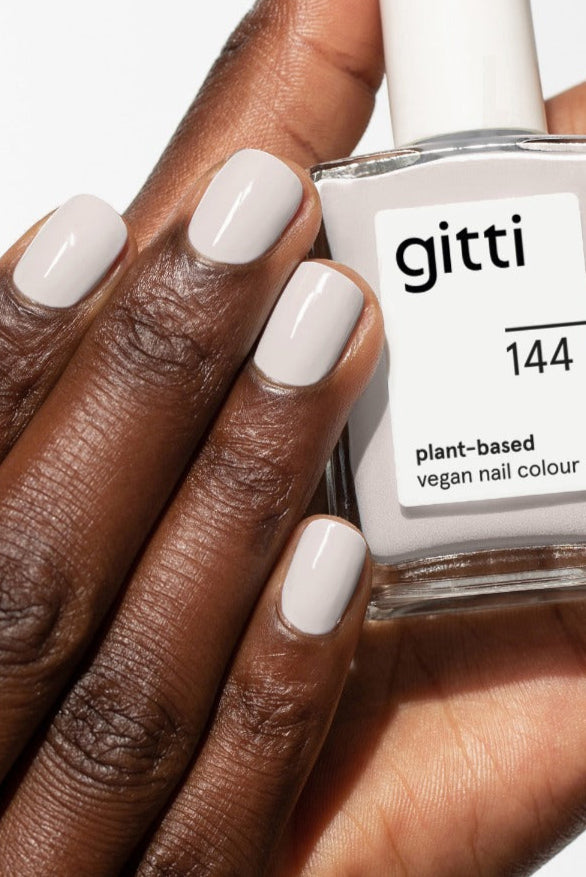 Eine Hand mit kurzen, manikürten Nägeln, die in einem zeitlosen Grau lackiert sind, hält eine Flasche mit gitti no. 144 pflanzlichen, veganen Nagellack.