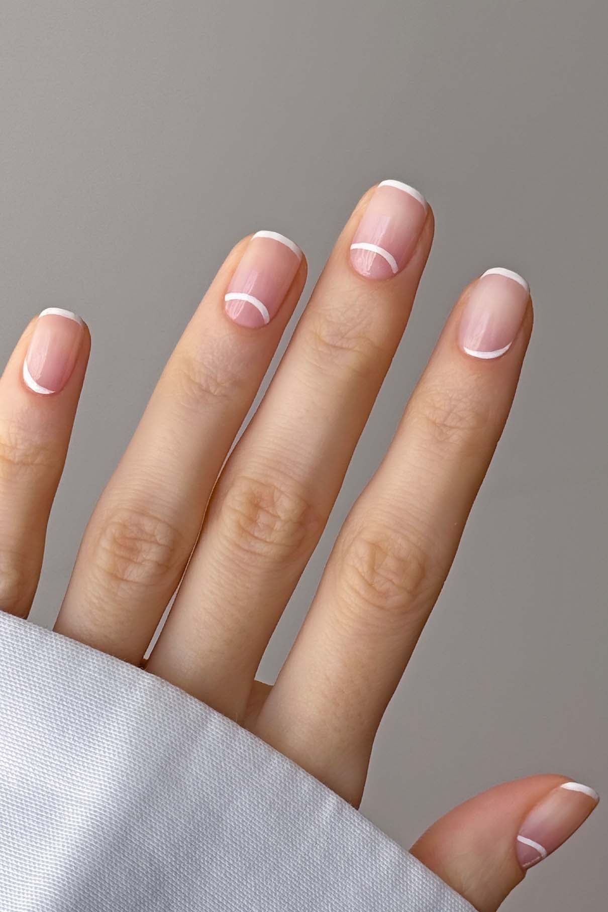 Eine Hand mit sauber manikürten Nägeln mit French Tip und weißem Halbkreisdesign in der Nähe der Nagelhaut, die mit Farben auf Pflanzenbasis aus dem Nail Art Set vor einem neutralen Hintergrund gestaltet wurden.