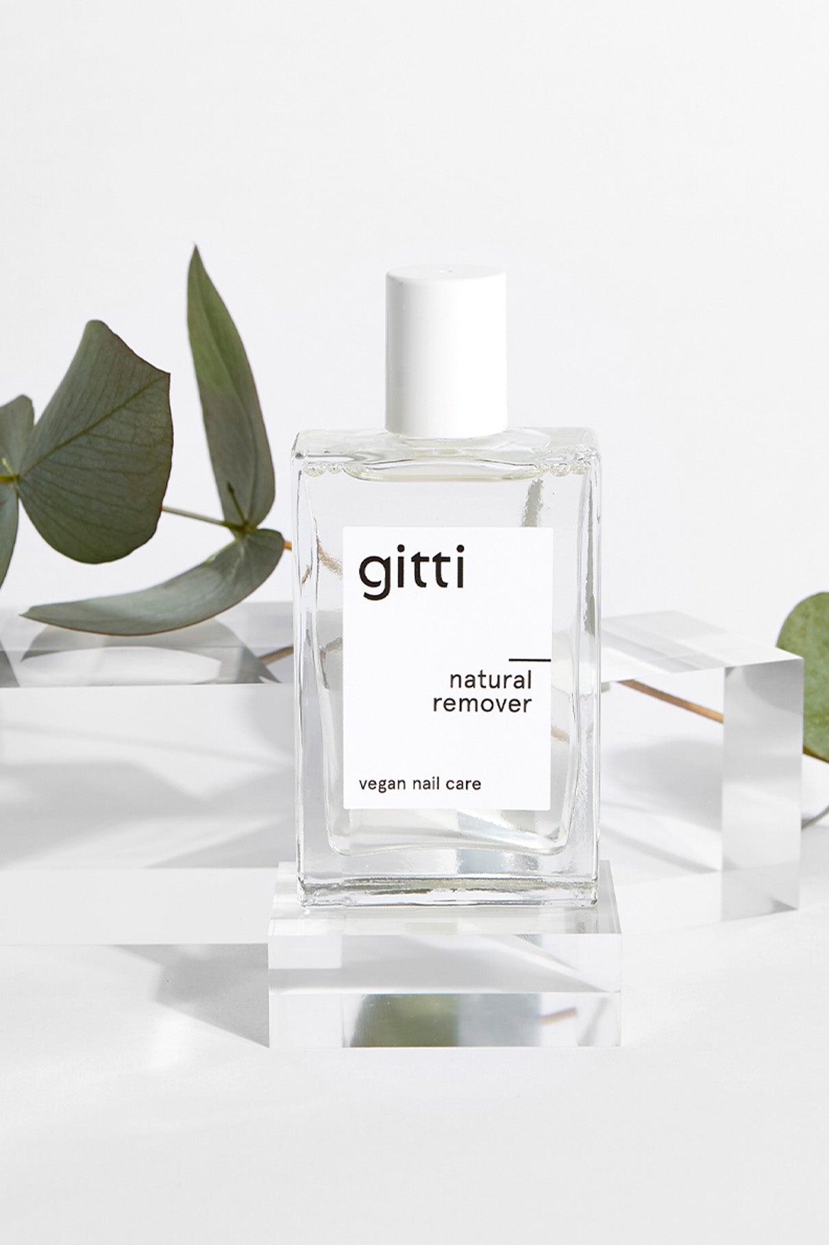 Eine Klarglasflasche mit der Aufschrift "gitti natural remover" und "vegane Nagelpflege" ruht auf einem transparenten Ständer neben grünen Eukalyptusblättern - ideal, um sie mit Ihrem Express All-in-One Set zu kombinieren.