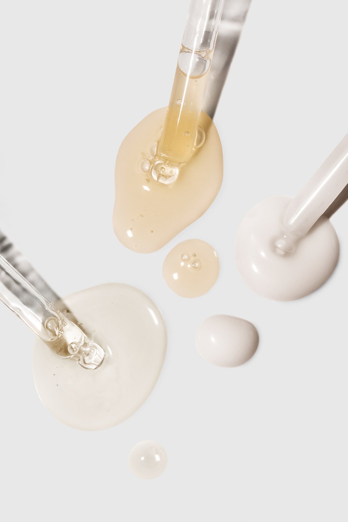 Zwei Pipetten aus dem Serum Duo spenden Pflegeseren in verschiedenen Farben, die kleine Pfützen und Tropfen auf einer weißen Fläche bilden.