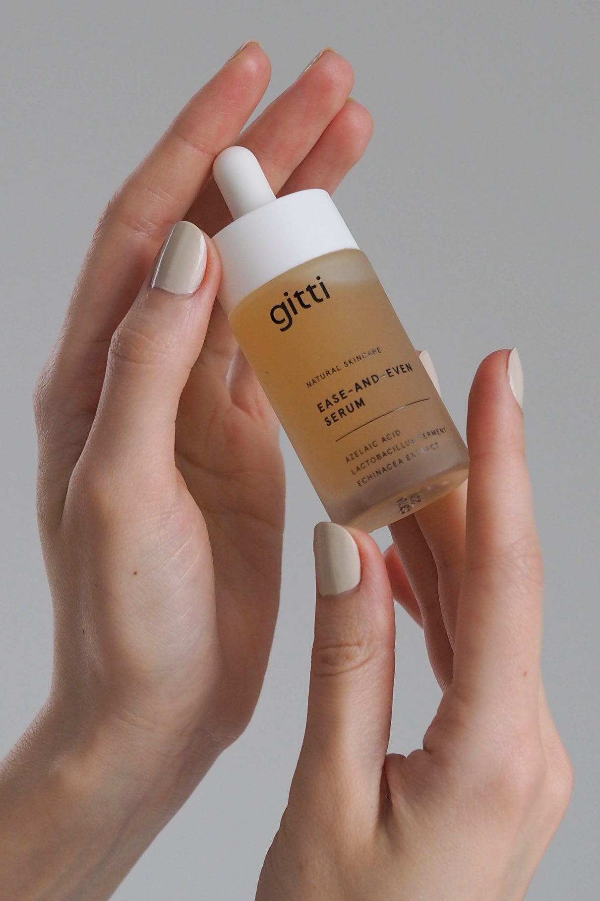Eine Hand mit hellem Nagellack hält vor einem neutralen Hintergrund ein Tropffläschchen mit der Aufschrift Ease-and-Even Serum, das Hautunreinheiten reduzieren und die Talgproduktion ausgleichen soll.